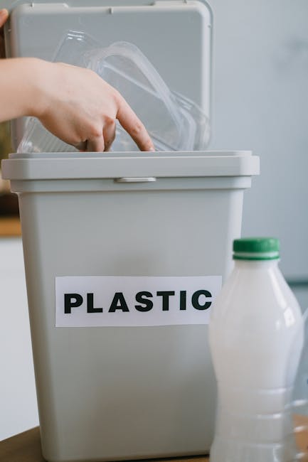 A hand is seen placing a clear plastic container into a grey recycling bin labeled 'PLASTIC', positioned on a wooden surface. In the foreground, there is a white plastic bottle with a green cap, placed beside the bin. The background appears to be an indoor setting with a light-colored wall, suggesting an environment focused on domestic waste sorting. The recycling bin's label clearly indicates it is designated for plastics, aligning with practices of separate waste collection, which can be part of private waste handling services offered by companies like Recycle Now. The scene emphasizes proper segregation of recyclable materials, reflecting common steps in responsible rubbish management and environmentally-conscious disposal routines inside households. The lighting is natural and soft, highlighting the textures of the plastic objects and the smooth surface of the bin. This visual supports the theme of rubbish removal and sustainable waste practices, focusing specifically on plastic waste sorting typical in alternative waste handling scenarios outside traditional local authority collections.