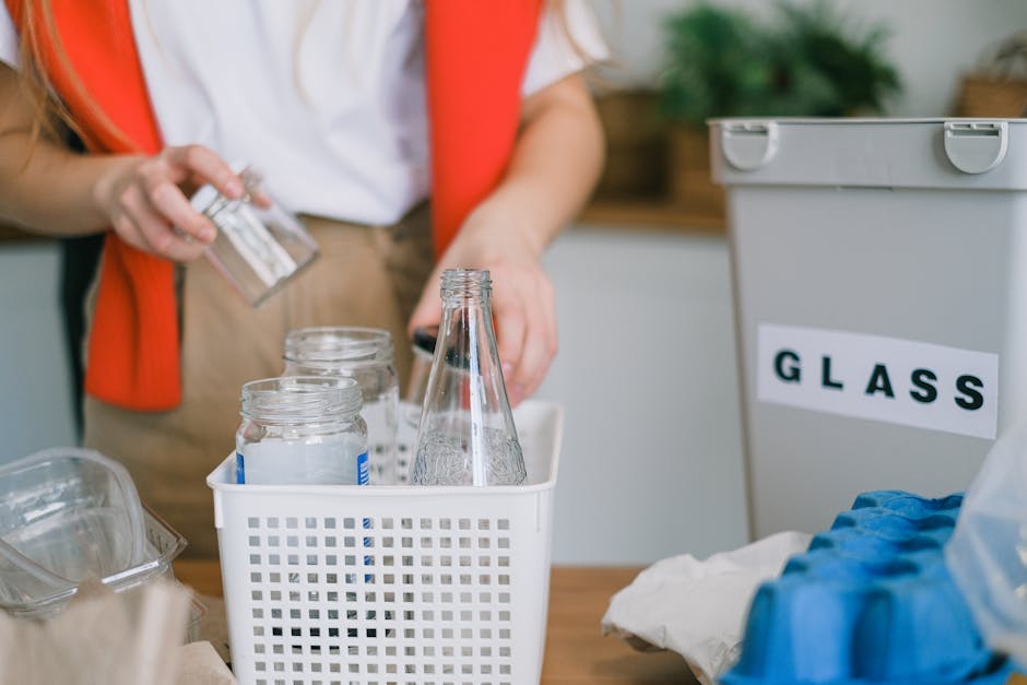 A hand is seen placing a clear plastic container into a grey recycling bin labeled 'PLASTIC', positioned on a wooden surface. In the foreground, there is a white plastic bottle with a green cap, placed beside the bin. The background appears to be an indoor setting with a light-colored wall, suggesting an environment focused on domestic waste sorting. The recycling bin's label clearly indicates it is designated for plastics, aligning with practices of separate waste collection, which can be part of private waste handling services offered by companies like Recycle Now. The scene emphasizes proper segregation of recyclable materials, reflecting common steps in responsible rubbish management and environmentally-conscious disposal routines inside households. The lighting is natural and soft, highlighting the textures of the plastic objects and the smooth surface of the bin. This visual supports the theme of rubbish removal and sustainable waste practices, focusing specifically on plastic waste sorting typical in alternative waste handling scenarios outside traditional local authority collections.