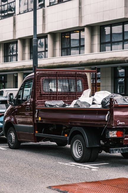 A small, dark red flatbed utility vehicle parked on a city street in front of a modern building with large glass windows and a concrete façade. The vehicle's open cargo area is filled with various types of waste, including large white and black plastic bags, loose debris, and possibly discarded materials. The bed is partially clear on the front side, with some items secured or leaning against the red metal side panel. The vehicle features a protective red metal grid at the cab's rear to prevent cargo from shifting into the driver’s area. The surroundings include pavement with a marked parking space, a streetlamp nearby, and additional parked vehicles visible in the background. The scene suggests an instance of private waste collection or on-site clearance, with the vehicle belonging to a rubbish removal service such as Recycle Now. The lighting indicates daytime, with natural daylight illuminating the scene, emphasizing the functional nature of the scene related to waste management and alternative rubbish handling methods outside of standard municipal collection.