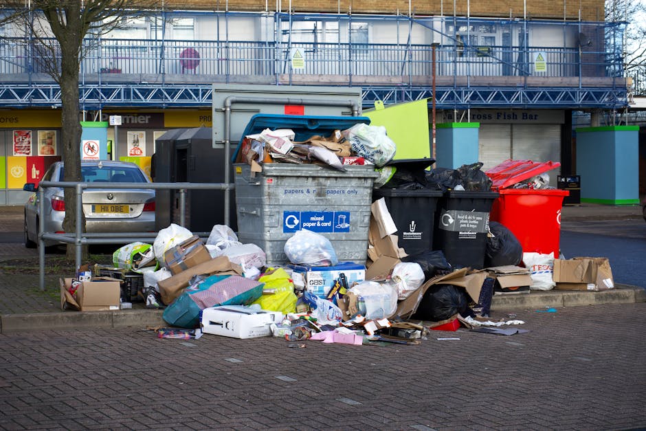 A collection of overflowing rubbish and waste materials piled against a metal railing on a paved urban street, including black, green, and red trash bins with some lids open, revealing mixed waste such as paper, cardboard boxes, plastic bags, and packaging materials. The waste spills onto the sidewalk, with some items on the ground, like crumpled paper, plastic containers, and discarded packaging, indicating a lack of proper disposal. In the background, there is a multi-story building with scaffolding and safety barriers, along with a row of commercial storefronts, one of which appears to be a fish bar. A parked car is situated beside the rubbish pile, partially blocking the sidewalk. The scene is well-lit with natural daytime light, emphasizing the cluttered and untidy state of the waste collection point, which may relate to private or alternative waste handling services, such as those provided by Recycle Now, servicing rubbish removal and on-site clearance needs.
