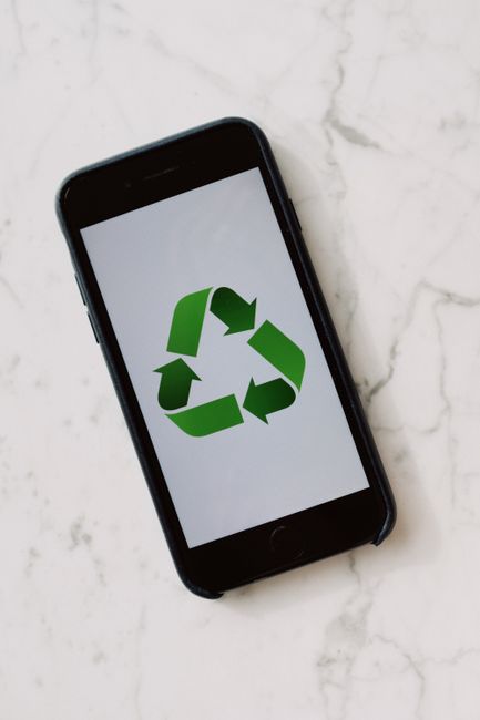 A black smartphone with rounded edges and a glossy finish is lying on a white marble surface. The phone's screen displays a large green recycling symbol composed of three arrows forming a triangle, with a matte finish that contrasts with the shiny screen. The marble surface has subtle grey veining and a polished texture, reflecting a soft, diffuse light. In the background, part of the marble surface is visible, suggesting a clean and minimalist environment. The image emphasizes the use of digital tools in sustainable waste management and the importance of electronics recycling, which companies like Recycle Now support through appropriate rubbish disposal services. The scene subtly highlights the theme of electronic waste, appropriate disposal methods, and private collection options, aligning with best practices in electronics recycling and data wiping within the context of responsible rubbish removal.